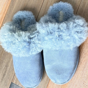 May include: A pair of light blue suede slippers featuring a plush, curly sheepskin lining and collar. The slip-on footwear has a rounded toe design and a visible brand label stitched onto the inner heel area of the soft, fuzzy interior lining.