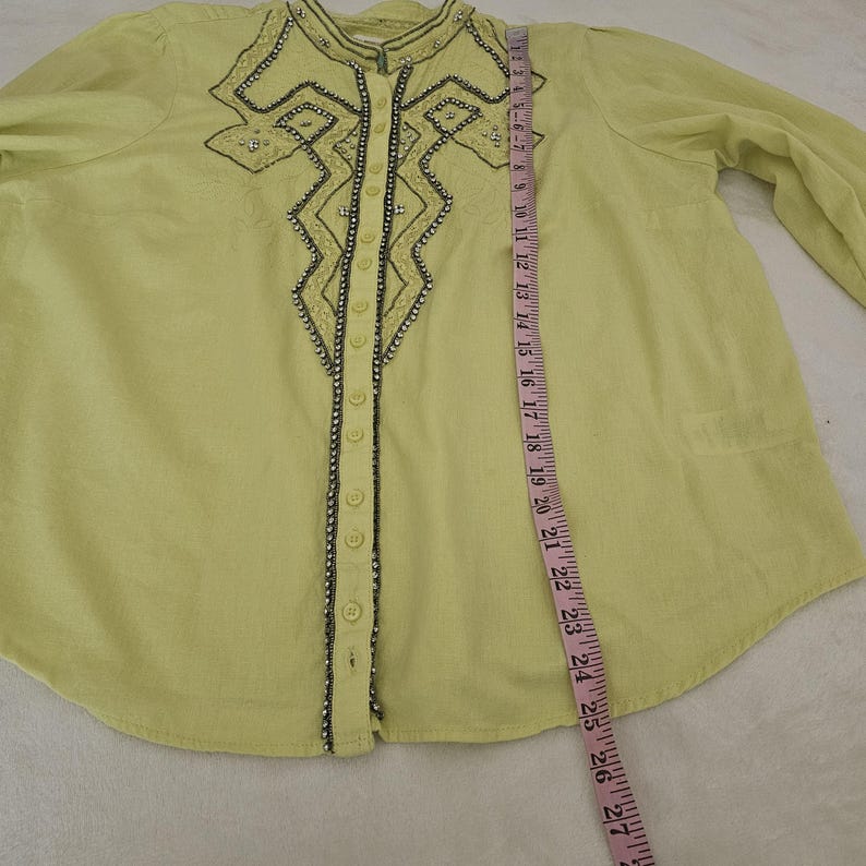 May include: A light green button-down shirt with long sleeves. The shirt features decorative beading and embroidery around the collar and down the front. A measuring tape shows the shirt is approximately 27 inches long.