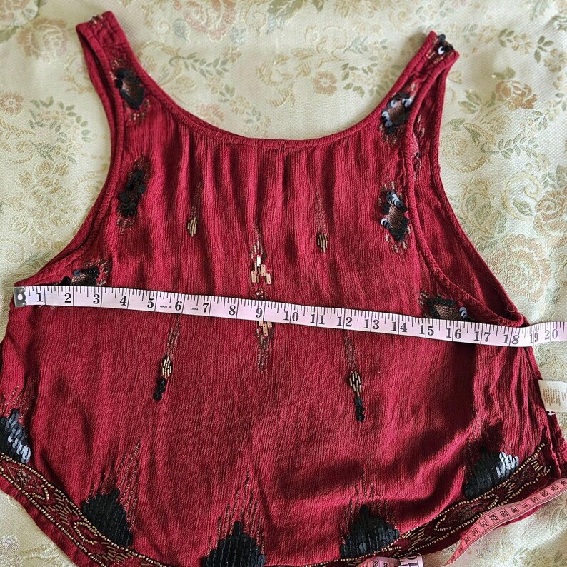 May include: A sleeveless, burgundy top with decorative beading and sequin accents. The top is measured with a tape measure, showing a width of approximately 45 cm. The fabric has a textured appearance.