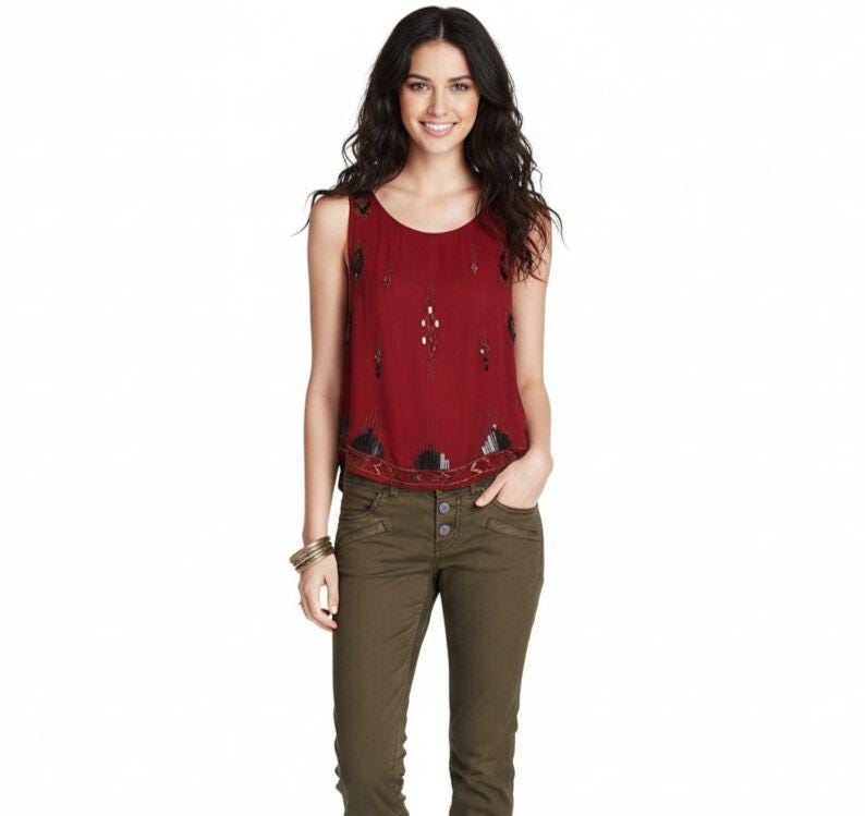May include: A sleeveless burgundy top with decorative beading and embellishments. The top is paired with olive green trousers. The model is wearing a gold bracelet.