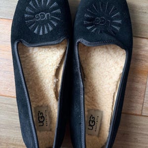 May include: A pair of black suede slip-on shoes featuring a sunburst-style UGG logo embroidered on the top. The interior is lined with beige shearling, and a white UGG Australia brand label is visible on the heel of the footbed.