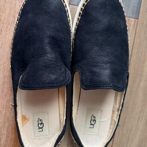 May include: A pair of black slip-on shoes with a smooth leather upper and a beige jute-wrapped midsole featuring visible stitching. The interior footbed is light beige and displays the UGG brand logo printed in black on the heel area.