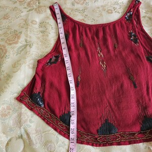 May include: A sleeveless, red top with decorative beading and embellishments. A pink measuring tape is placed vertically along the garment, showing measurements up to 56 cm. The top has a scoop neckline and a flared hemline.