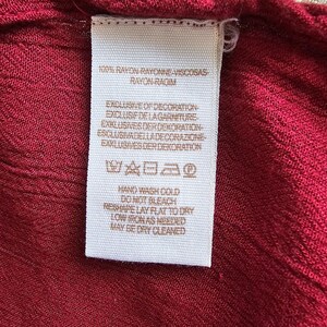 May include: A white clothing label with black text against a red fabric. The label provides care instructions and fabric information, including "RAYON" and "VISCOSAS". The fabric is a deep red colour.