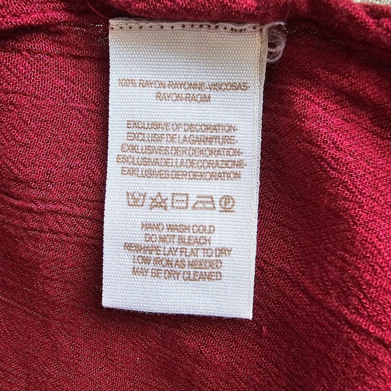 May include: A white clothing label with black text against a red fabric. The label provides care instructions and fabric information, including "RAYON" and "VISCOSAS". The fabric is a deep red colour.