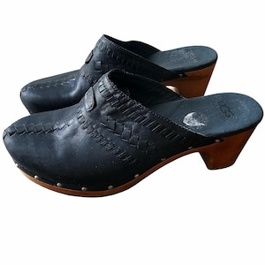 May include: A pair of black leather clogs with a woven design on the vamp. The clogs have wooden heels and soles, with silver-colored studs around the base. The brand name "UGG" is visible on the insole.