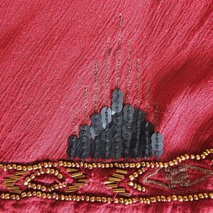 May include: Close-up of a red fabric with a textured surface. The fabric is embellished with black sequins in a tiered design and gold beads along the bottom edge. Gold chain-like accents are also present.