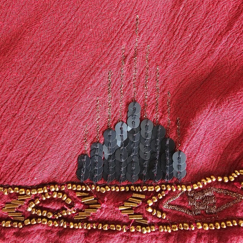 May include: Close-up of a red fabric with a textured surface. The fabric is embellished with black sequins in a tiered design and gold beads along the bottom edge. Gold chain-like accents are also present.