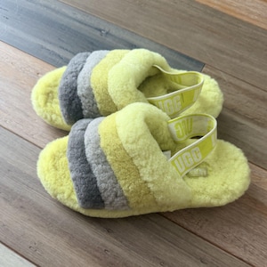 May include: A pair of fuzzy slippers featuring a multi-colored design with bands of bright yellow, light gray, and dark gray sheepskin. Each slipper includes a yellow elastic heel strap with the white text UGG printed repeatedly along the band.