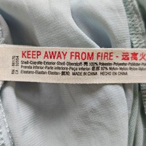 Może przedstawiać: Zbliżenie jasnoniebieskiej tkaniny z białą metką. Na metce znajduje się czerwony napis "KEEP AWAY FROM FIRE" oraz chińskie znaki. Na metce podano r&oacute;wnież skład tkaniny: 100% poliester, 92% nylon i elastan. Wyprodukowano w Chinach.