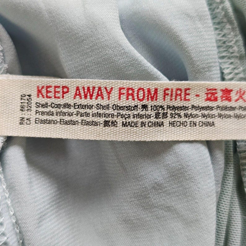Może przedstawiać: Zbliżenie jasnoniebieskiej tkaniny z białą metką. Na metce znajduje się czerwony napis "KEEP AWAY FROM FIRE" oraz chińskie znaki. Na metce podano r&oacute;wnież skład tkaniny: 100% poliester, 92% nylon i elastan. Wyprodukowano w Chinach.