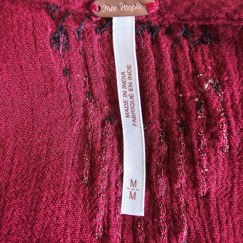 May include: Close-up of a burgundy fabric with a textured surface. The image shows a tag with the brand name "Free People" and a label that reads "MADE IN INDIA FABRIQU&Eacute; EN INDE". The tag also has the size "M".