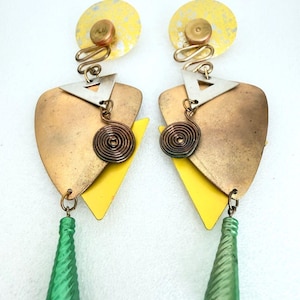 May include: A pair of dangle earrings featuring a yellow circular top, a silver triangle, a bronze-toned shield-shaped plate, a dark metal spiral, and a green twisted cone-shaped bead. The design incorporates layered geometric shapes and mixed metal finishes.
