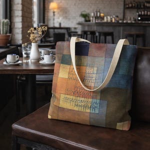 Pode incluir: Uma sacola colorida com um design geométrico em tons de laranja, azul e marrom. A sacola possui alças creme e é exibida em um banco de couro marrom em um ambiente de café. A obra de arte na sacola parece ser um design arquitetônico abstrato.