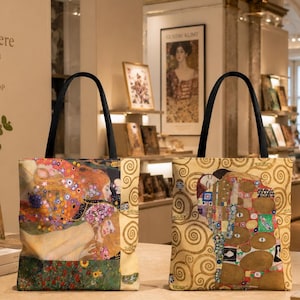 Puede incluir: Dos bolsos tote rectangulares con asas negras, cada uno con un estampado colorido de una obra de Gustav Klimt. El bolso de la izquierda muestra una escena con figuras y flores, mientras que el de la derecha representa el icónico abrazo dorado de El Beso.