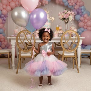 Puede incluir: Una niña con un vestido plateado y blanco brillante de un solo hombro con un lazo rosa, sosteniendo globos plateados, morados y rosas. El fondo presenta un ambiente de fiesta con globos, una mesa y sillas doradas.
