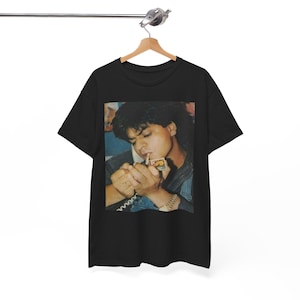 Puede incluir: Camiseta negra con una imagen vintage de una persona encendiendo un cigarrillo. La imagen es un primer plano de la cara y las manos de la persona, con una chaqueta vaquera visible. La camiseta está colgada de una percha de madera.