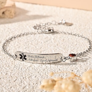 May include: A silver medical alert bracelet with a chain and a rectangular plate. The plate has a black medical symbol and engraved text: "Boston Bellamah, Naloxone - Opioid Antagonist." A small, round, orange gemstone is set near the clasp.