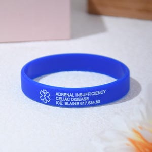 May include: A vibrant blue medical alert bracelet. The bracelet features white text that reads "ADRENAL INSUFFICIENCY CELIAC DISEASE ICE: ELAINE 617.834.80" and a medical symbol. The bracelet is made of a flexible material.