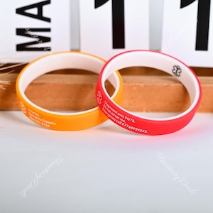 May include: Two silicone wristbands with white inner bands and contrasting outer bands. One is orange with the text "Ananda Diabetic" and a phone number. The other is red with the text "Diagnosed with POTS. Do not stress me. Emergency call 07768939545."