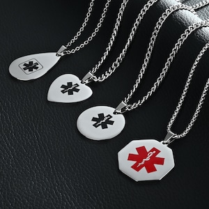 Può includere: Una collezione di collane di allerta medica argentate con varie forme di ciondolo: a goccia, a cuore e rotondo. Ogni ciondolo presenta un simbolo medico nero. Una collana ha un simbolo medico rosso su un ciondolo ottagonale. Le collane sono su una superficie nera.