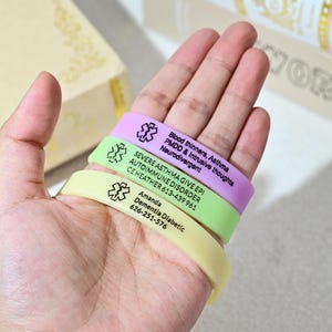 May include: Three medical alert bracelets in pastel shades: lilac, lime green, and pale yellow. Each bracelet features a medical symbol and printed text detailing medical conditions and contact details. The bracelets are held in a hand.