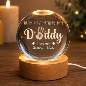 Puede incluir: Una esfera de cristal transparente con las palabras "HAPPY FIRST FATHER'S DAY!" y "Daddy" grabadas, junto con una huella de mano y el mensaje "I love you Jessica - 2026". La esfera descansa sobre una base de madera iluminada.