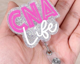 May include: A badge reel with the text "CNA Life" in pink and white. The letters "CNA" are in a bold, blocky font, while "Life" is in a cursive script. The background is silver glitter. The reel has a clear plastic strap and a metal clip.