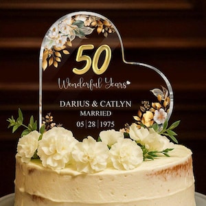 May include: A clear heart-shaped acrylic cake topper featuring the gold number 50, the text "Wonderful Years", "DARIUS & CATLYN", "MARRIED", and "05 | 28 | 1975". The topper is decorated with gold and white floral illustrations and sits on a frosted cake.