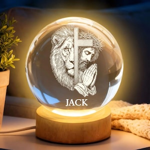 May include: A clear glass sphere featuring a detailed engraving of a lion's head and a figure with a cross, illuminated from below. It sits on a wooden base, with the name "JACK" inscribed. The warm light enhances the intricate design.