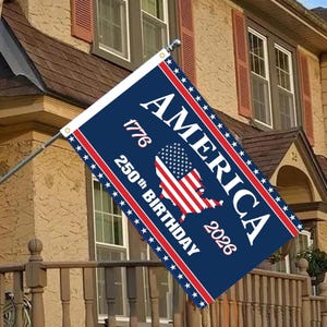 May include: A rectangular flag with a dark blue background, featuring the word "AMERICA" in white capital letters. The flag includes the years "1776" and "2026", and the phrase "250th BIRTHDAY". The flag also displays a map of the United States with the American flag design.
