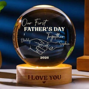 Puede incluir: Una esfera de cristal transparente con la inscripción "Our First Father's Day Together" y los nombres "Daddy" y "Sophia". La esfera descansa sobre una base de madera que dice "I LOVE YOU". También se muestra el año "2026".