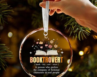 Könnte beinhalten: Ein klarer, runder Weihnachtsschmuck mit weißem Band. Der Schmuck zeigt das Wort "BOOKTROVERT" über einem offenen Buch mit Blumen und Schmetterlingen. Der Text darunter definiert einen Bücherwurm.