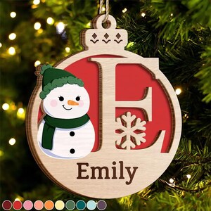 May include: Wooden Christmas ornament with a snowman and the letter "E" inside a red circle. The ornament has the name "Emily" at the bottom and a snowflake design. The snowman wears a green hat and scarf.