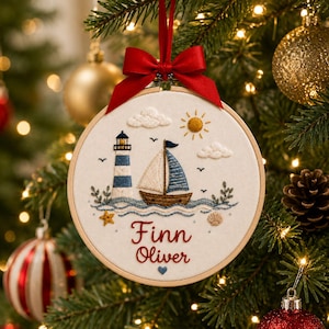 May include: A circular embroidery hoop ornament featuring a stitched lighthouse, a sailboat, clouds, a sun, a starfish, and a seashell on off-white fabric. The name Finn Oliver is embroidered in red script below the scene with a small blue heart.