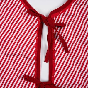 May include: A quilted vest featuring a red and white diagonal striped pattern. The garment has a round neckline and a front opening secured by two sets of red fabric ties. The edges are finished with solid red binding, and the quilted texture.