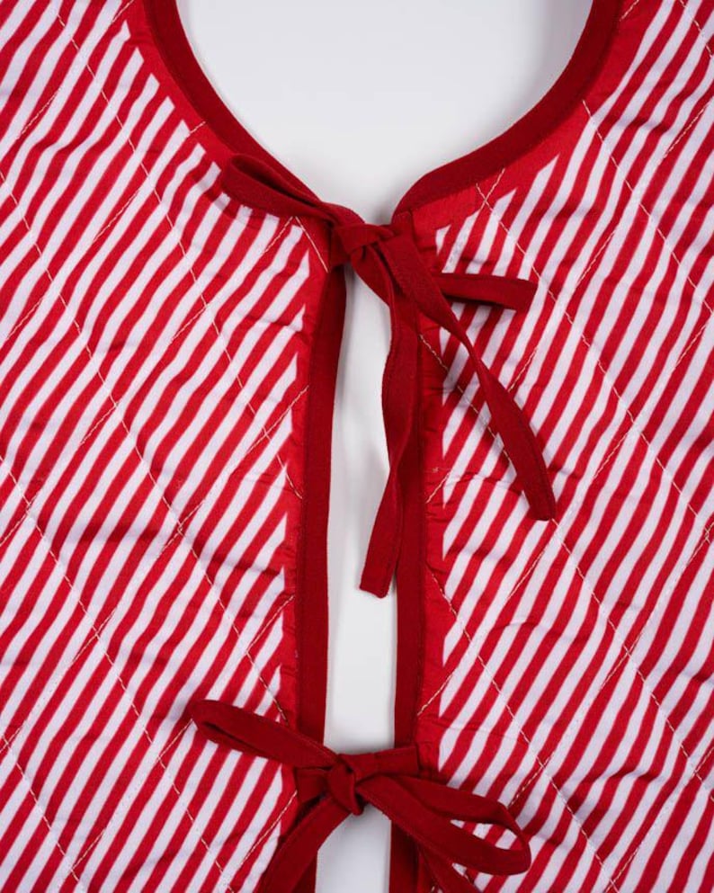 May include: A quilted vest featuring a red and white diagonal striped pattern. The garment has a round neckline and a front opening secured by two sets of red fabric ties. The edges are finished with solid red binding, and the quilted texture.