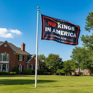 May include: A black flag with red trim and white text reading "NO KINGS IN AMERICA SINCE 1776" is displayed on a flagpole. The flag waves in the wind against a backdrop of a blue sky, green grass, and a brick house.