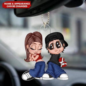 May include: A car charm featuring a cartoon couple. The woman wears a red top, blue jeans, and hoop earrings. The man wears a black shirt, black cap, and blue jeans, holding a heart-shaped gift. The text "NAME & APPEARANCE CAN BE CHANGED" is at the top.