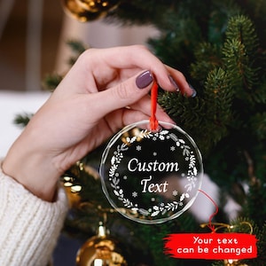 May include: A clear, round Christmas ornament with a red ribbon, held by a person. The ornament has a black background with white floral and snowflake designs, and the words "Custom Text". The text "Your text can be changed" is in a red rectangle.