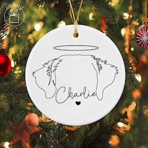 May include: White ceramic Christmas ornament with a black line drawing of a dog's head, a halo, and the name "Charlie" with a heart. The ornament hangs from a gold string against a blurred Christmas tree background.