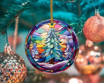 Puede incluir: Adorno navideño redondo con un diseño de vidriera que presenta un árbol de Navidad estilizado, nieve y una colorida puesta de sol. El adorno está bordeado en morado y tiene un cordón dorado para colgar. Otros adornos y un árbol de Navidad están en el fondo.