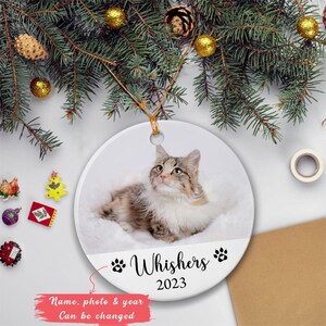 以下が含まれることがあります： 猫の写真が入った丸い白いセラミック製のクリスマスオーナメント。「Whishers 2023」の文字と足跡がプリントされています。金色の紐で吊り下げられており、背景にはクリスマスの飾りがあります。