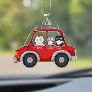 May include: A red car-shaped ornament with three cartoon cats inside. The cats are white, gray, and black, each with a unique expression. The names "Lucci", "Max", and "Mimi" are printed below each cat. The ornament hangs from a silver chain.