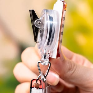 May include: A clear plastic retractable badge reel with a black metal clip and a decorative rectangular badge. The badge has a red, white, and yellow design. The reel is attached to a silver-colored metal clasp.