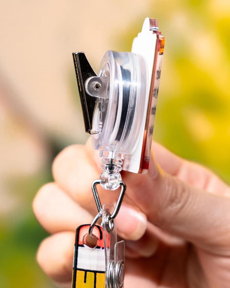 May include: A clear plastic retractable badge reel with a black metal clip and a decorative rectangular badge. The badge has a red, white, and yellow design. The reel is attached to a silver-colored metal clasp.