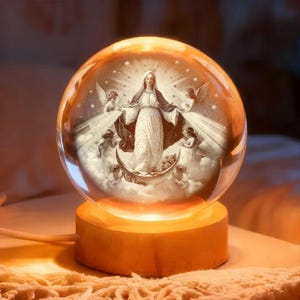 May include: A clear glass sphere with an image of a religious figure surrounded by angels, set on a wooden base. The sphere is illuminated from within, creating a warm, glowing effect. The image inside depicts a woman in a white gown with a dark cloak.
