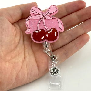 Op de afbeelding: Een roze intrekbare badge reel met een kersen- en strikdesign. De badge reel heeft een roze glitterachtergrond, een lichtroze strik en twee rode kersen. De reel heeft een doorzichtige plastic houder en een metalen clip.
