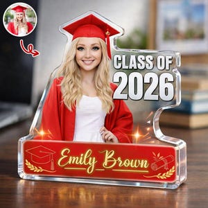 Könnte beinhalten: Ein personalisiertes Abschlussandenken mit einem Foto einer lächelnden Absolventin in roter Kappe und Robe. Das klare Acrylteil trägt den Text "CLASS OF 2026" und den Namen "Emily Brown" in goldfarbenen Buchstaben, zusammen mit Abschluss-Symbolen.