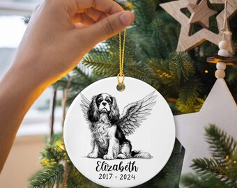May include: A round, white Christmas ornament featuring a black and white Cavalier King Charles Spaniel with angel wings. The ornament has a gold string for hanging and includes the name "Elizabeth" and the years "2017-2024".